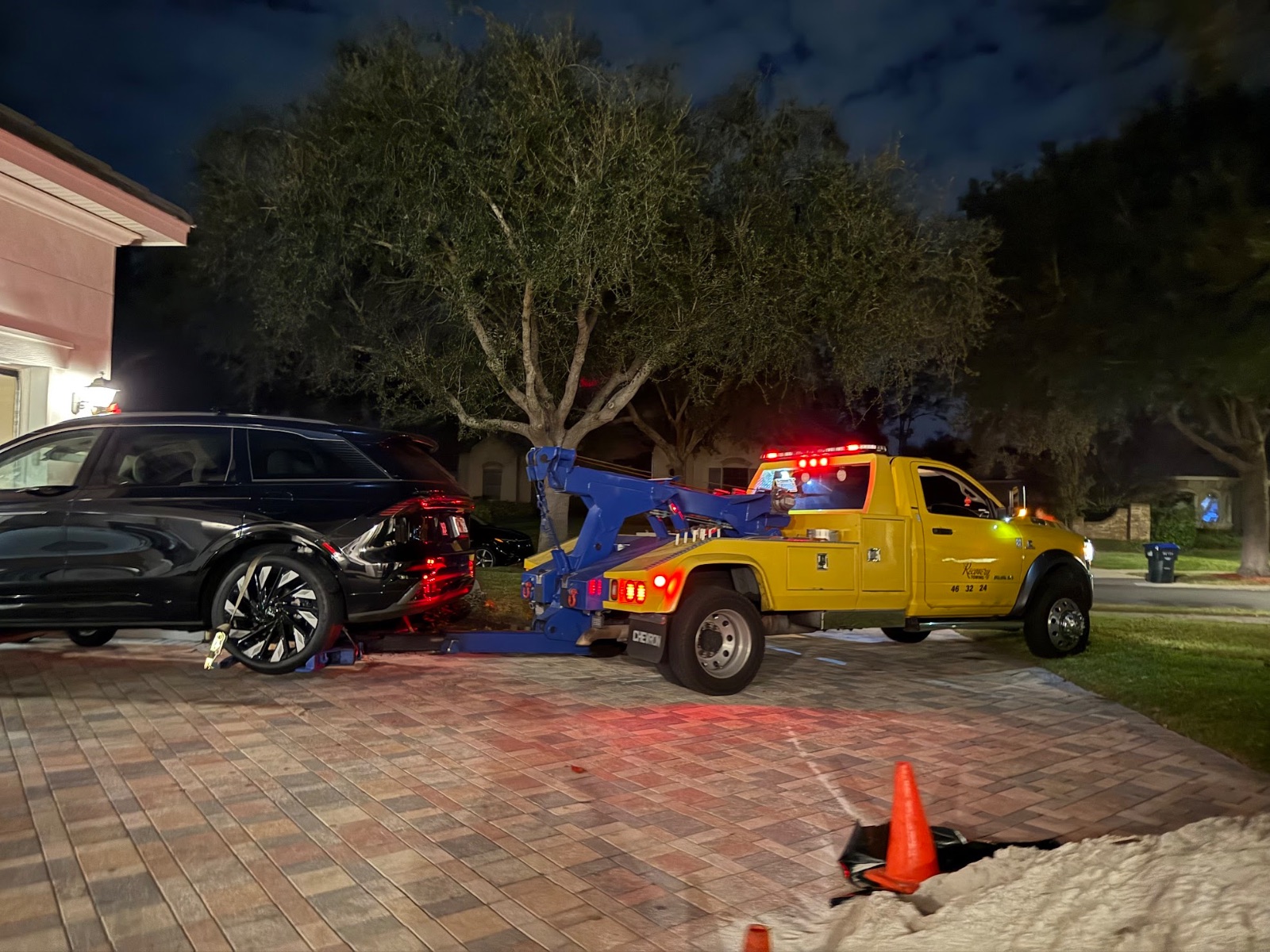 Yellow tow truck with blue crane lifting damaged black car at night in residential driveway