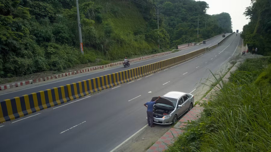 Driver inspecting engine of silver sedan on highway shoulder with yellow safety barrier